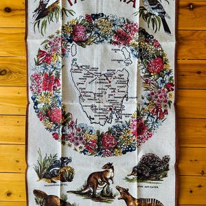 Puede incluir: Una toalla de cocina blanca con un borde floral colorido y un mapa de Tasmania en el centro. El mapa est&aacute; rodeado de ilustraciones de animales nativos de Tasmania, incluyendo un kookaburra, un p&aacute;jaro wattle, un demonio de Tasmania, un wallaby, un hormiguero espinoso, un ornitorrinco y un tigre de Tasmania. El texto "TASMANIA" est&aacute; impreso en rojo en la parte superior de la toalla. El texto "DESIGNED BY TASILE" est&aacute; impreso en la parte inferior de la toalla.