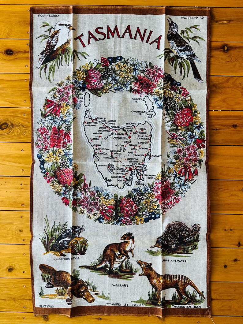 Puede incluir: Una toalla de cocina blanca con un borde floral colorido y un mapa de Tasmania en el centro. El mapa est&aacute; rodeado de ilustraciones de animales nativos de Tasmania, incluyendo un kookaburra, un p&aacute;jaro wattle, un demonio de Tasmania, un wallaby, un hormiguero espinoso, un ornitorrinco y un tigre de Tasmania. El texto "TASMANIA" est&aacute; impreso en rojo en la parte superior de la toalla. El texto "DESIGNED BY TASILE" est&aacute; impreso en la parte inferior de la toalla.