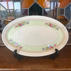 May include: Oval serving platter with a light green centre and floral accents. The rim features a delicate, openwork design. The floral pattern includes pink, blue, and yellow flowers. The platter is displayed on a black stand.