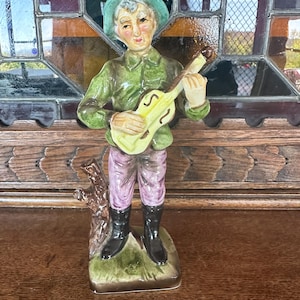 Puede incluir: Una figurita de cerámica de una persona tocando la guitarra. La figurita está pintada en tonos de verde, marrón, amarillo y rosa. La persona lleva un sombrero verde y tiene barba blanca. La figurita está de pie sobre una base marrón.