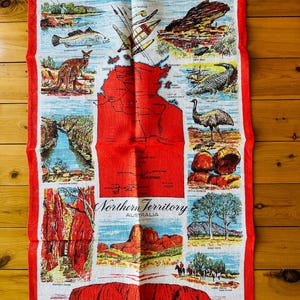 Op de afbeelding: Een rode en witte linnen theedoek met een kaart van het Northern Territory van Australi&euml;. De kaart is omringd door afbeeldingen van verschillende bezienswaardigheden en dieren uit de regio, waaronder Uluru, een krokodil, een emoe en een kangoeroe. De tekst "Northern Territory Australia" is in zwart gedrukt aan de onderkant van de theedoek.