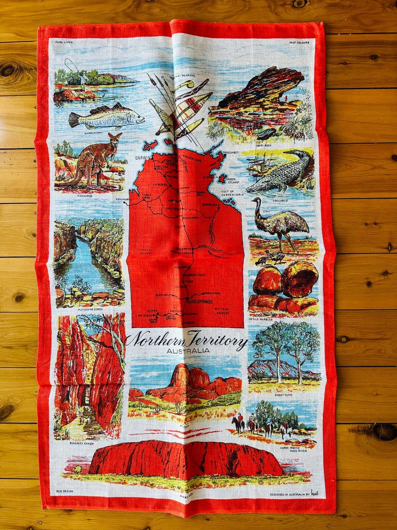 Op de afbeelding: Een rode en witte linnen theedoek met een kaart van het Northern Territory van Australi&euml;. De kaart is omringd door afbeeldingen van verschillende bezienswaardigheden en dieren uit de regio, waaronder Uluru, een krokodil, een emoe en een kangoeroe. De tekst "Northern Territory Australia" is in zwart gedrukt aan de onderkant van de theedoek.