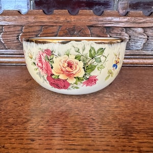 May include: A white ceramic bowl with a gold rim and a floral design featuring pink, yellow, and red roses. The bowl is sitting on a wooden surface.