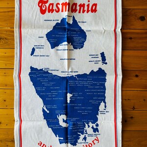 Puede incluir: Una toalla de cocina blanca con un mapa azul y rojo de Tasmania y el texto "Tasmania y su territorio". El mapa est&aacute; lleno de texto que describe las caracter&iacute;sticas &uacute;nicas de la isla, como "El aire m&aacute;s limpio del mundo", "La mejor cerveza del mundo" y "El sitio hist&oacute;rico m&aacute;s grande del mundo".