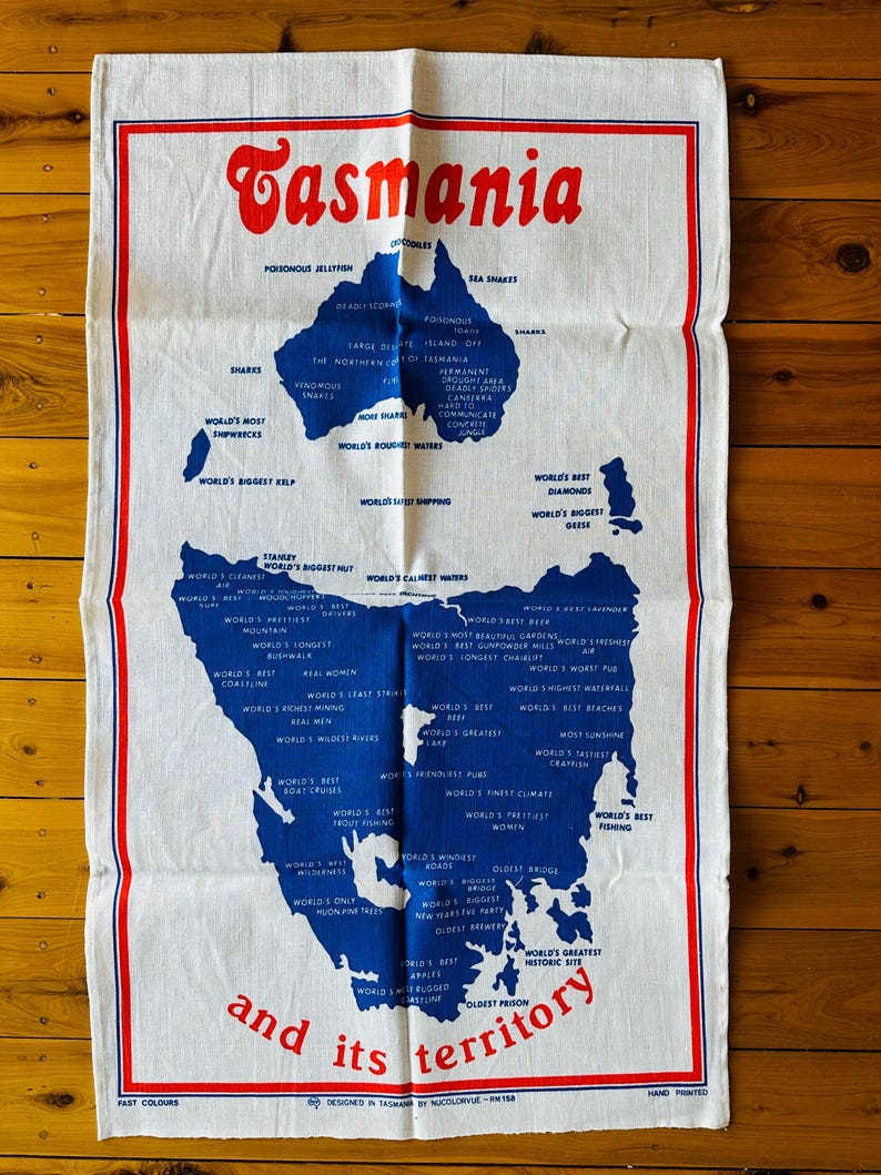 Puede incluir: Una toalla de cocina blanca con un mapa azul y rojo de Tasmania y el texto "Tasmania y su territorio". El mapa est&aacute; lleno de texto que describe las caracter&iacute;sticas &uacute;nicas de la isla, como "El aire m&aacute;s limpio del mundo", "La mejor cerveza del mundo" y "El sitio hist&oacute;rico m&aacute;s grande del mundo".