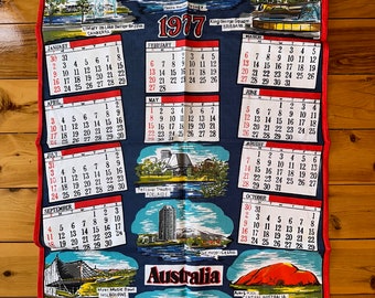 Op de afbeelding: Een blauwe en rode theedoek met een kalender voor het jaar 1977. De kalender bevat afbeeldingen van bezienswaardigheden uit Australië, waaronder het Sydney Opera House, de Harbour Bridge en Ayers Rock. De tekst "Australië" staat in grote letters onderaan de theedoek.