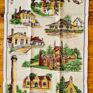 Puede incluir: Un pa&ntilde;o de cocina vintage con fondo beige e ilustraciones coloridas de edificios hist&oacute;ricos en Tasmania. El texto "Historic Tasmania" est&aacute; impreso en letras marrones en la parte superior de la toalla. La toalla presenta ilustraciones de edificios como el puente de Richmond, Port Cottage, Emily House, Arthur Circus, East Tower, Penny Royal y Backland Church.