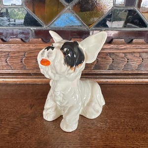 May include: A white ceramic figurine of a small dog with black and brown markings on its face. The dog is sitting with its head tilted up and its mouth slightly open.