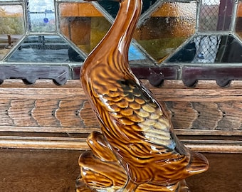 May include: A brown ceramic duck figurine with a long neck and detailed feather textures. The duck is perched on a base that resembles a log. The figurine has a glossy finish and is likely a decorative item.