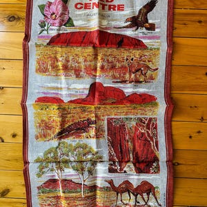 Op de afbeelding: Een vintage linnen theedoek met een kleurrijke illustratie van het Rode Centrum van Australi&euml;. Het ontwerp omvat Uluru, de Ghost Gums, een wilde kameel, een dingo, een stekelduivelhagedis en een wigstaart arend. De tekst "RED CENTRE AUSTRALIA" is in rood gedrukt.