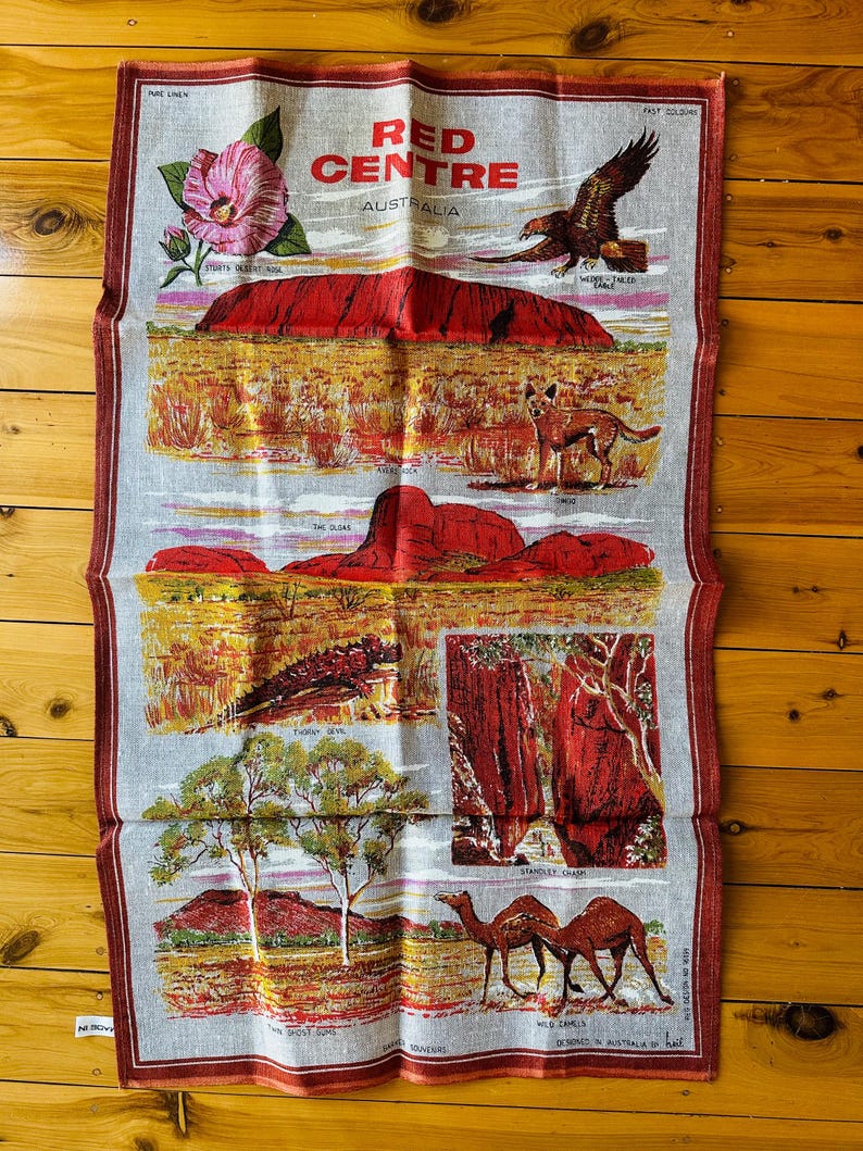 Op de afbeelding: Een vintage linnen theedoek met een kleurrijke illustratie van het Rode Centrum van Australi&euml;. Het ontwerp omvat Uluru, de Ghost Gums, een wilde kameel, een dingo, een stekelduivelhagedis en een wigstaart arend. De tekst "RED CENTRE AUSTRALIA" is in rood gedrukt.