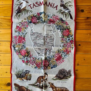 Puede incluir: Una toalla de t&eacute; de lino con un mapa de Tasmania, Australia, rodeado de una corona de flores nativas. El mapa incluye las principales ciudades y pueblos. La parte inferior de la toalla presenta ilustraciones de animales nativos, incluyendo un ornitorrinco, un demonio de Tasmania, un ualab&iacute;, un tigre de Tasmania y un equidna.