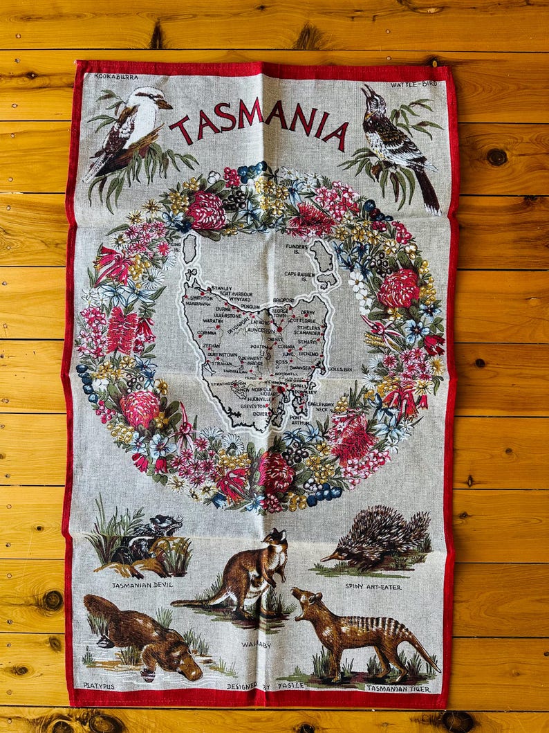 Puede incluir: Una toalla de t&eacute; de lino con un mapa de Tasmania, Australia, rodeado de una corona de flores nativas. El mapa incluye las principales ciudades y pueblos. La parte inferior de la toalla presenta ilustraciones de animales nativos, incluyendo un ornitorrinco, un demonio de Tasmania, un ualab&iacute;, un tigre de Tasmania y un equidna.