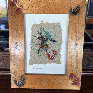 May include: A framed artwork featuring a Native American-inspired painting of a figure in a red and black outfit, holding a flute. The painting is on textured paper with red handprints and a blue and green background. The frame is wood with black handprints on the corners.