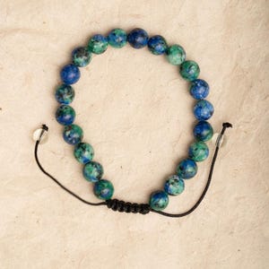 Pode incluir: Pulseira de contas redondas e polidas em tons de azul e verde. As contas são enfiadas em um cordão preto com extremidades ajustáveis. A pulseira é exibida em uma superfície marrom clara.