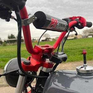 Könnte beinhalten: Nahaufnahme eines roten Motorradlenkers mit einem schwarzen Schaumstoffgriff und einem schwarzen und silbernen Tachometer. Der Lenker ist an einem schwarzen Motorrad mit einem silbernen Tank montiert.