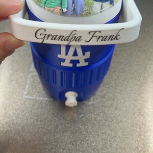 K&ouml;nnte beinhalten: Blau-wei&szlig;e K&uuml;hlbox mit einem Foto auf dem Deckel. Der Deckel zeigt ein Bild eines Mannes und zweier Kinder. Auf der K&uuml;hlbox steht "Grandpa Frank" und ein wei&szlig;es "LA"-Logo.