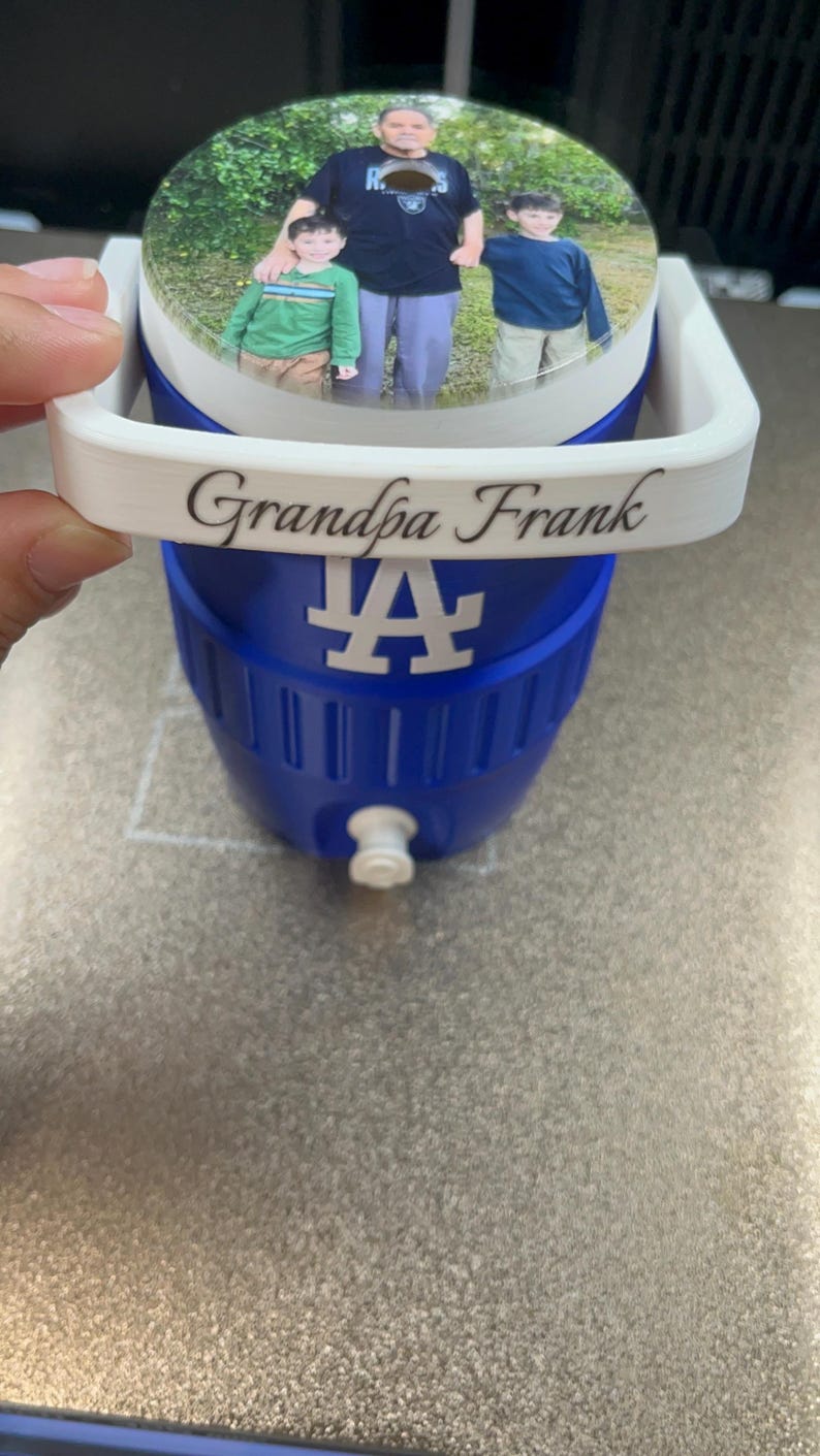 K&ouml;nnte beinhalten: Blau-wei&szlig;e K&uuml;hlbox mit einem Foto auf dem Deckel. Der Deckel zeigt ein Bild eines Mannes und zweier Kinder. Auf der K&uuml;hlbox steht "Grandpa Frank" und ein wei&szlig;es "LA"-Logo.