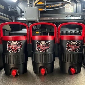 K&ouml;nnte beinhalten: Drei schwarze Wasserk&uuml;hler mit roten Deckeln und Griffen. Jeder K&uuml;hler hat einen roten Wasserhahn und einen Aufkleber mit der Aufschrift "Azteca Market" mit einem Baseballschl&auml;ger- und Ball-Design. Die K&uuml;hler stehen auf einer grauen Oberfl&auml;che.