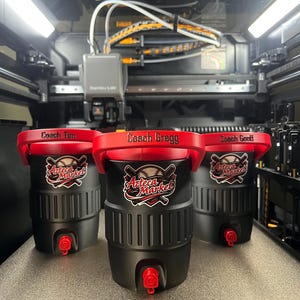 K&ouml;nnte beinhalten: Drei schwarze und rote Wasserk&uuml;hler mit den Texten "Coach Tim", "Coach Gregg" und "Coach Geoff" auf den Deckeln. Jeder K&uuml;hler hat das "Azteca Market" Logo und einen roten Wasserhahn. Die K&uuml;hler stehen auf einem 3D-Drucker.