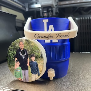 K&ouml;nnte beinhalten: Ein blauer Wasserk&uuml;hler mit wei&szlig;em Griff und dem Text "Grandpa Frank". Der K&uuml;hler hat das Logo der Los Angeles Dodgers. Ein runder Untersetzer mit einem Foto eines Mannes und zweier Kinder befindet sich neben dem K&uuml;hler.