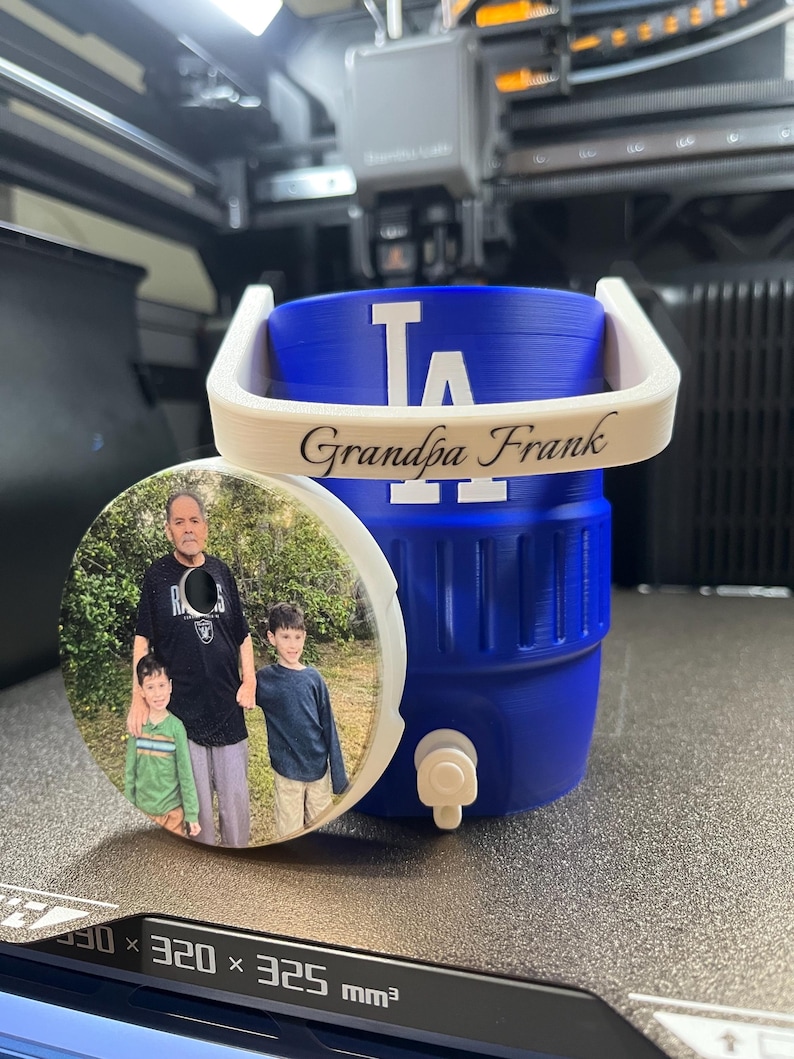 K&ouml;nnte beinhalten: Ein blauer Wasserk&uuml;hler mit wei&szlig;em Griff und dem Text "Grandpa Frank". Der K&uuml;hler hat das Logo der Los Angeles Dodgers. Ein runder Untersetzer mit einem Foto eines Mannes und zweier Kinder befindet sich neben dem K&uuml;hler.