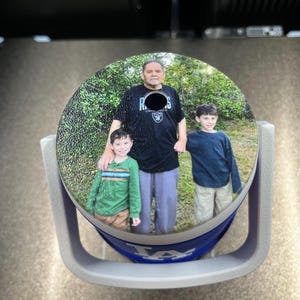 K&ouml;nnte beinhalten: Ein runder Auto-Untersetzer mit einem Familienfoto. Der Untersetzer befindet sich in einem grauen Halter mit blauem und wei&szlig;em Design. Das Foto zeigt einen Mann und zwei Jungen vor einem gr&uuml;nen Hintergrund.