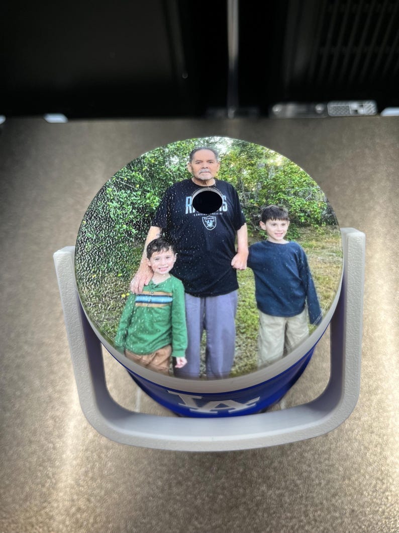 K&ouml;nnte beinhalten: Ein runder Auto-Untersetzer mit einem Familienfoto. Der Untersetzer befindet sich in einem grauen Halter mit blauem und wei&szlig;em Design. Das Foto zeigt einen Mann und zwei Jungen vor einem gr&uuml;nen Hintergrund.