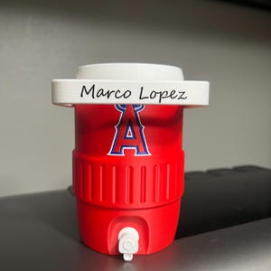K&ouml;nnte beinhalten: Ein roter Wasserk&uuml;hler mit wei&szlig;em Deckel und wei&szlig;em Wasserhahn. Der K&uuml;hler zeigt das Logo der Los Angeles Angels in Blau und Wei&szlig;. Der Name "Marco Lopez" ist auf der Oberseite aufgedruckt.