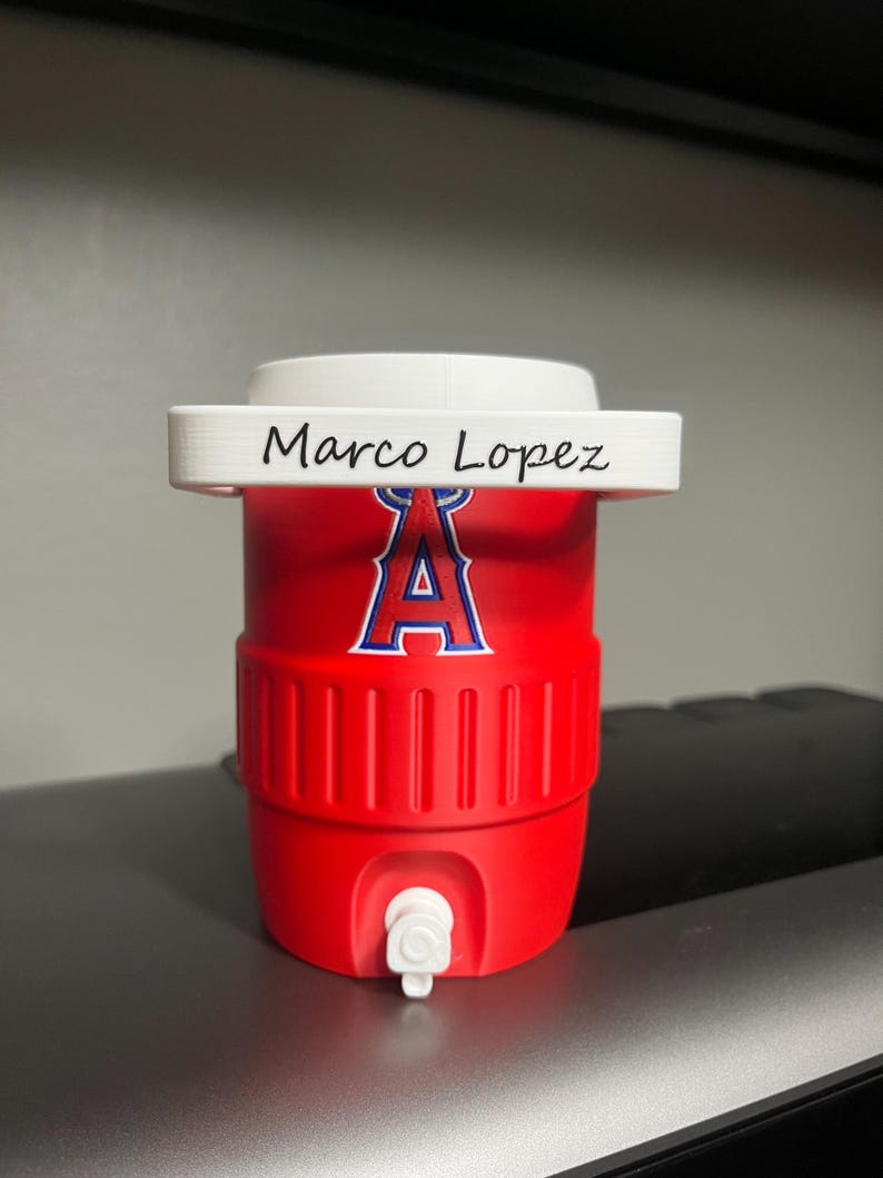 K&ouml;nnte beinhalten: Ein roter Wasserk&uuml;hler mit wei&szlig;em Deckel und wei&szlig;em Wasserhahn. Der K&uuml;hler zeigt das Logo der Los Angeles Angels in Blau und Wei&szlig;. Der Name "Marco Lopez" ist auf der Oberseite aufgedruckt.