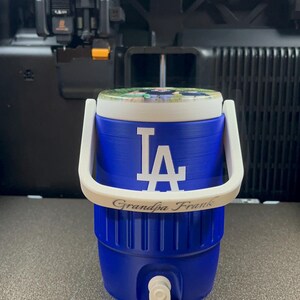 K&ouml;nnte beinhalten: Ein blauer Wasserk&uuml;hler mit wei&szlig;em Griff und Deckel. Der K&uuml;hler zeigt die wei&szlig;en Buchstaben "LA" und den Text "Grandpa Frank". Der K&uuml;hler hat einen wei&szlig;en Wasserhahn und wird wahrscheinlich verwendet, um Getr&auml;nke kalt zu halten.