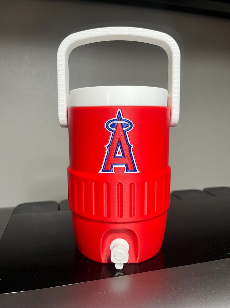 K&ouml;nnte beinhalten: Ein roter Wasserk&uuml;hler mit wei&szlig;em Griff und Deckel. Der K&uuml;hler zeigt das Logo der Los Angeles Angels in Blau und Wei&szlig;. Er hat einen wei&szlig;en Wasserhahn an der Unterseite zum Ausgeben von Fl&uuml;ssigkeiten. Der K&uuml;hler ist so konzipiert, dass Getr&auml;nke kalt gehalten werden.
