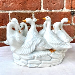 May include: A white ceramic figurine featuring a group of geese arranged in a circle, each with a yellow beak and orange feet. The geese are sculpted with detailed feather textures and are positioned on a base molded to resemble a stone nest.