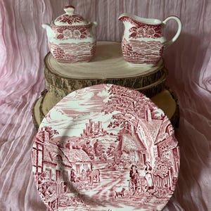 May include: A red and white ceramic tea set with a vintage design. The set comprises a lidded sugar bowl, a creamer jug, and a large plate. The plate showcases a detailed rural scene. The items are presented on a wooden surface against a pink background.
