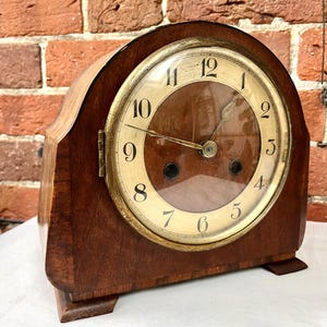 May include: A wooden mantel clock with a curved top and a circular face. The clock features a cream-coloured dial with black Arabic numerals, gold-toned hands, and a brass-coloured bezel. Two winding holes are visible on the lower half of the dial.