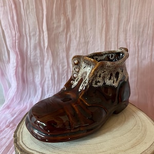 Puede incluir: Una maceta de cerámica marrón con forma de bota y acabado brillante. La bota tiene un diseño detallado con un ribete texturizado de color claro alrededor de la parte superior y ojales. Se encuentra sobre una rodaja de madera sobre un fondo de tela rosa.