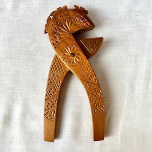 May include: A light brown wooden nutcracker featuring intricate hand-carved geometric patterns, including star-shaped motifs and cross-hatch designs along the two handles and the curved upper section, connected by a central metal pivot pin.