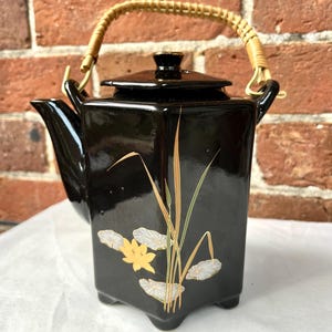 May include: A glossy black ceramic teapot with a hexagonal body and a removable lid. The teapot features a painted design of a yellow flower and green reeds. It has a curved spout and a light brown woven bamboo handle attached to two metal loops.