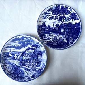 May include: Two round ceramic plates featuring blue and white transferware patterns. One plate depicts a horse-drawn carriage on a country road near a village. The other plate shows a large tree, a river, and a village scene with a church steeple.