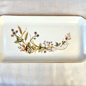 Puede incluir: Una bandeja de servir rectangular de cerámica color blanco hueso con bordes festoneados y un borde dorado. El centro presenta una ilustración botánica de tallos de trigo, hojas verdes, pequeñas flores blancas, moras y bayas rojas.