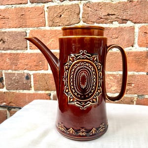 May include: A tall, cylindrical brown ceramic coffee pot with a glossy finish. The body features a central embossed oval medallion with intricate cream-coloured scrollwork and a matching decorative border around the base, a curved spout, and a handle.
