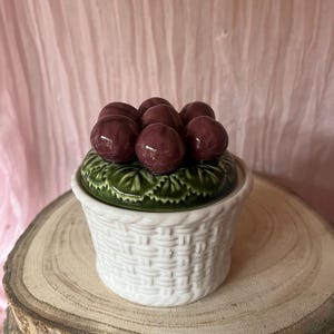 May include: A white ceramic basket-shaped container with a lid featuring a cluster of dark red, glossy spheres. The lid has a green base with a leaf design. The container sits on a wooden slice.