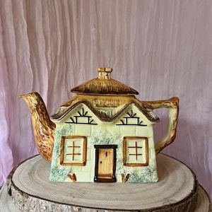 May include: A cottage-shaped ceramic teapot with a thatched roof and a light yellow facade, complete with windows and a door. The spout and handle are brown. The teapot is displayed on a wooden slice.