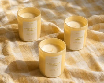 Peut inclure: Trois bougies cylindriques en verre remplies de cire jaune pâle. Chaque bougie présente une étiquette rectangulaire blanche avec le texte Limoncello Creme imprimé en lettres grasses, suivi d'un texte plus petit. Une mèche est centrée.