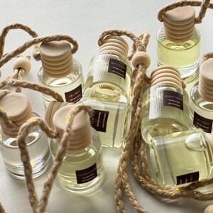 May include: Clear glass car air freshener bottles with wooden caps and beige rope hangers. The bottles are filled with a light yellow liquid and have small rectangular labels. The rope has gold metallic accents.