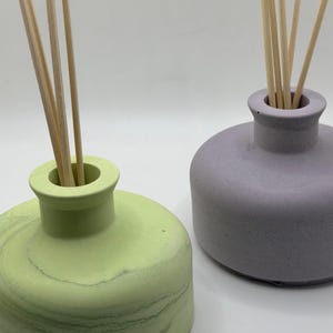 May include: Two decorative reed diffuser bottles. One is light green with a marbled effect, the other is lavender. Each has a narrow neck and contains several wooden reeds. The bottles are on a white background.