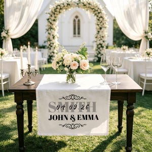 May include: A wooden table set for a wedding ceremony, with a white table runner featuring the names "Smith" and "John & Emma" along with the date "04.09.26". The table is decorated with a floral arrangement, candles, and wine glasses, set against a backdrop of a floral arch.