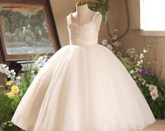 May include: A cream-colored formal dress on a mannequin. The bodice features a ruched design with delicate straps and embellishments. The full skirt is made of layers of tulle, creating a voluminous silhouette. The dress is displayed on a gold-colored stand.