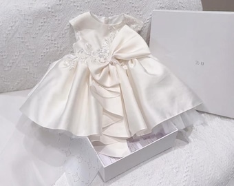 May include: Ivory satin baby dress with a large bow and delicate lace detailing. The dress has a full skirt and is presented in a white gift box. A beautiful outfit for a special occasion.