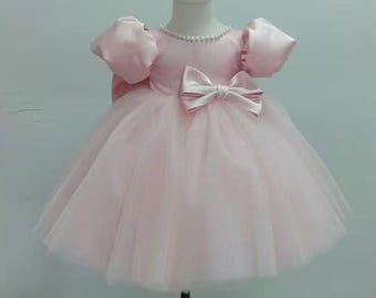 May include: A light pink baby dress with a tulle skirt and satin bodice. The dress features a bow at the waist, puff sleeves, and a pearl necklace. The dress is displayed on a mannequin.